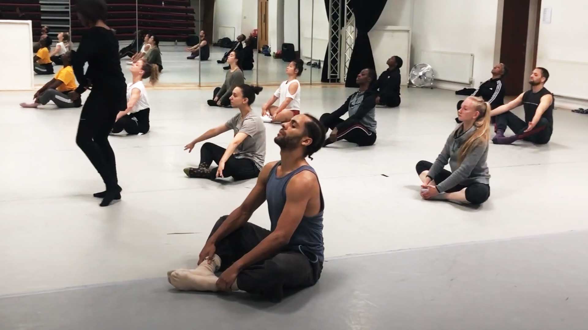 A group of dancers sitting on a dance studio floor with their legs crossed and head facing the celing